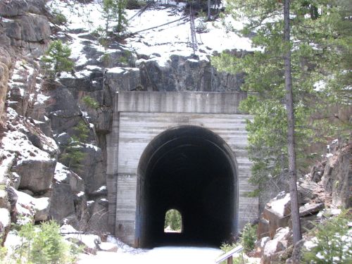 A dark tunnel entrance carved into rocky terrain, surrounded by trees and snow.