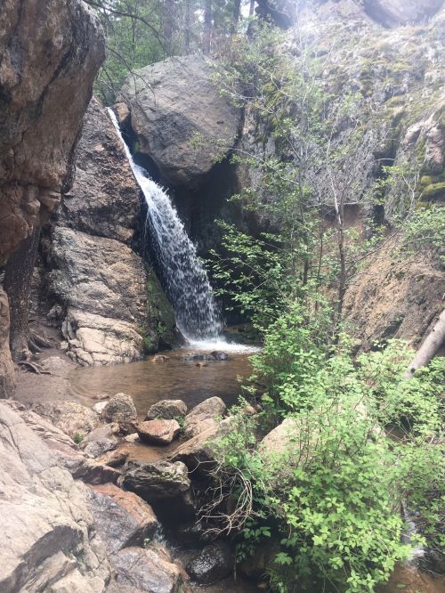 A serene waterfall cascades over rocks into a small pool, surrounded by lush greenery and rugged terrain.