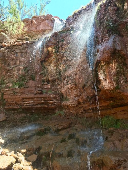 A waterfall cascading down a rocky cliff, surrounded by greenery and sunlight.