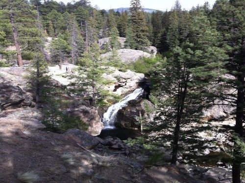A serene waterfall cascades over rocks, surrounded by tall pine trees and rugged terrain in a forested landscape.