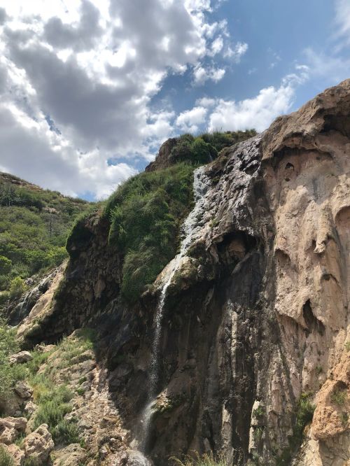 A waterfall cascading down a rocky cliff, surrounded by lush greenery and a blue sky with scattered clouds.