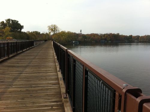 A wooden walkway extends over a calm river, surrounded by trees with autumn foliage.