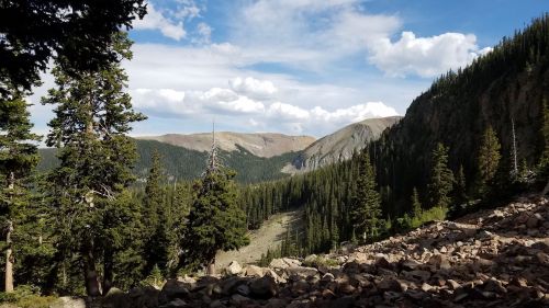 A scenic view of a mountainous landscape with pine trees, rocky terrain, and a blue sky with scattered clouds.
