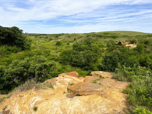 A scenic view of rolling green hills and rocky outcrops under a blue sky with wispy clouds.