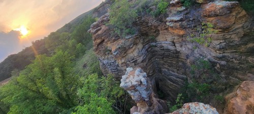 A rocky cliffside with layered stone formations, surrounded by lush greenery and a sunset in the background.