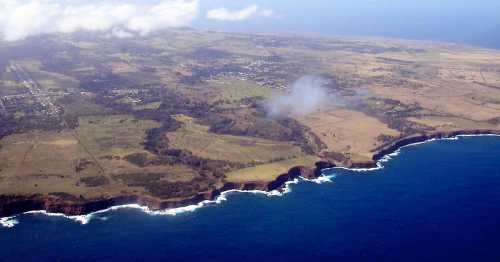 Aerial view of a coastal landscape with cliffs, greenery, and the ocean under a partly cloudy sky.