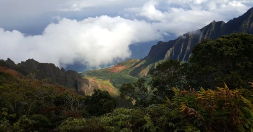 A scenic view of lush mountains and coastline, with clouds hovering over the landscape and the ocean in the distance.