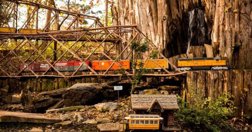 A miniature train set runs through a forest scene with a wooden bridge, rocks, and a small cabin near a waterfall.