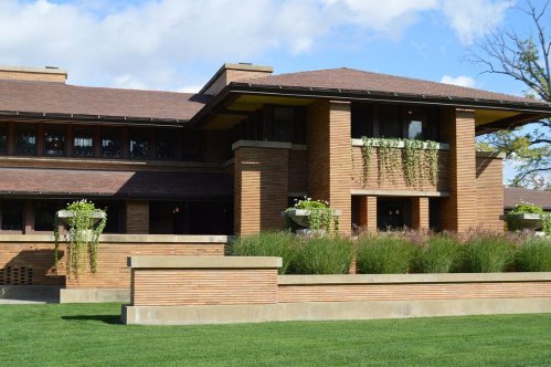 A modernist brick house with large windows, green planters, and a landscaped lawn under a blue sky.