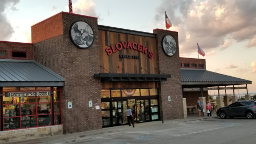 Exterior of Slovacek's store with wooden accents, flags, and a sign, during sunset. People are entering and exiting.