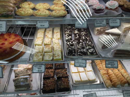 A display case filled with various desserts, including cupcakes, bars, and cakes, all labeled with names.