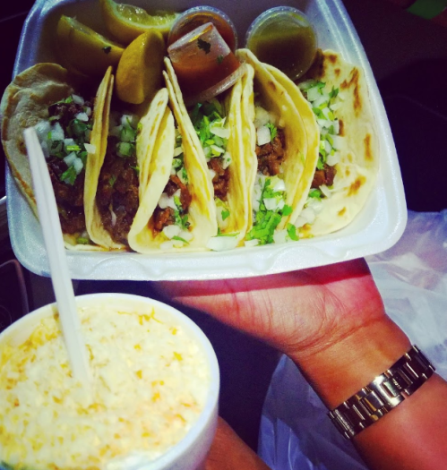 A hand holding a takeout container with four tacos topped with onions and cilantro, alongside a cup of corn.