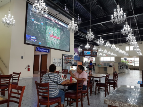 A spacious indoor area with chandeliers, featuring a couple sitting at a table and a large TV displaying a weather report.
