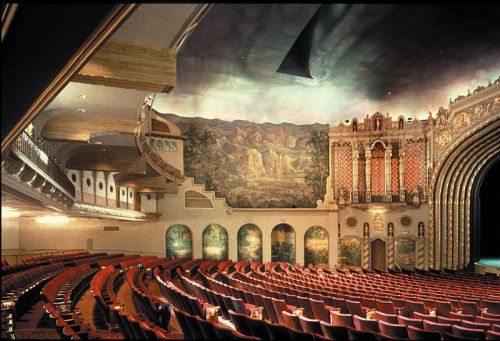 Interior of a vintage theater featuring ornate architecture, red seating, and a large mural on the stage backdrop.