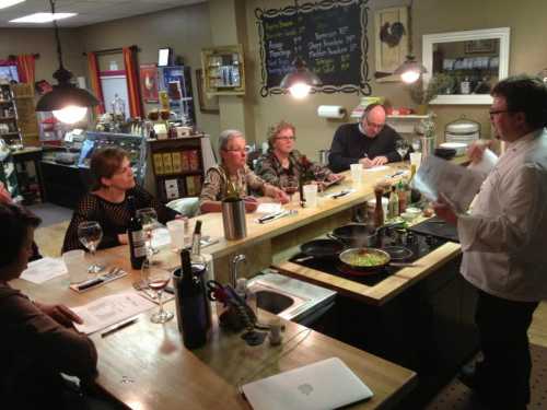 A chef demonstrates cooking techniques to a small group at a kitchen counter, with wine glasses and ingredients visible.