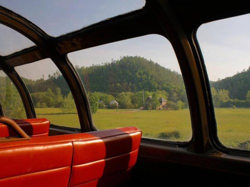 View from a train's observation car, showcasing lush green hills and a rural landscape through large windows.