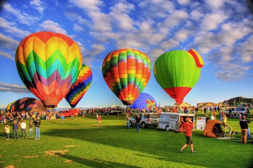 Colorful hot air balloons fill the sky at a lively festival, with crowds enjoying the vibrant scene on a sunny day.