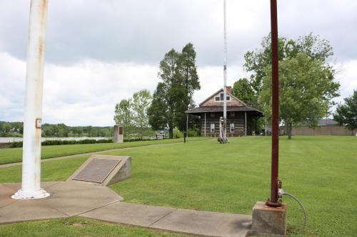 A grassy area with a historic building, trees, and a flagpole near a body of water under a cloudy sky.