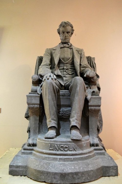 A seated statue of Abraham Lincoln, dressed in a suit, with a solemn expression, resting on a grand chair.