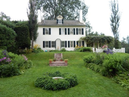 A charming white house with black shutters, surrounded by lush gardens and colorful flowers on a green lawn.