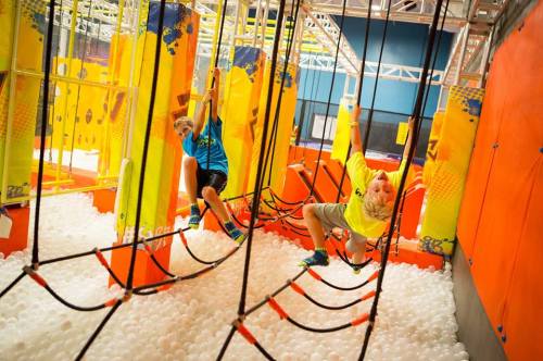 Two kids play on a colorful indoor obstacle course, swinging on ropes above a pit filled with white balls.