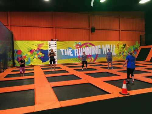 A vibrant indoor trampoline park with people jumping on orange trampolines and a colorful wall mural in the background.
