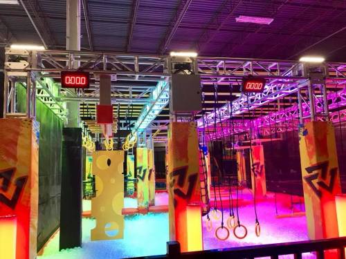 Colorful indoor obstacle course with various hanging rings and climbing structures, illuminated by vibrant lights.