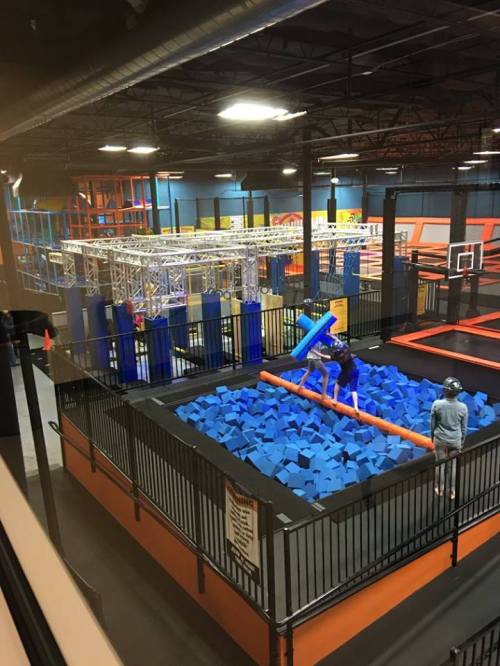 A view of a trampoline park with a foam pit, featuring blue foam blocks and people playing on a balance beam.