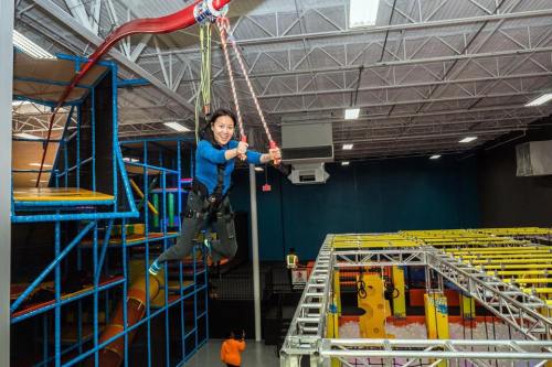 A person in a blue shirt swings on a zip line in an indoor adventure park, surrounded by colorful obstacle courses.