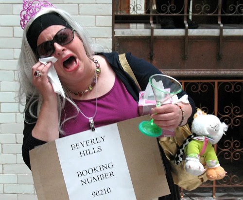 A woman in a tiara and gray wig, holding a drink and a sign, appears distressed while wiping her tears.
