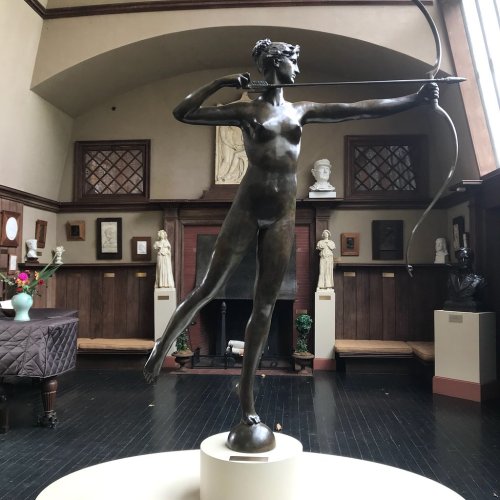 A bronze statue of a woman archer stands in a gallery, surrounded by artworks and sculptures in a well-lit room.