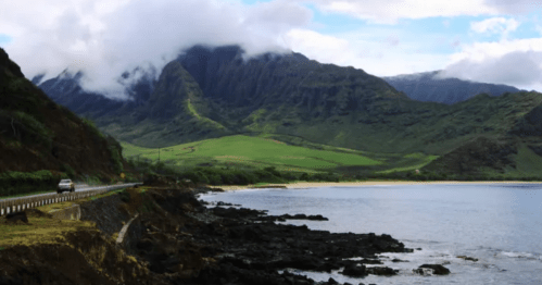 A scenic coastal road winds along rocky shores, with lush green mountains and clouds in the background.