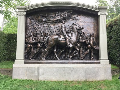 A bronze relief sculpture depicting a cavalry leader and soldiers marching, surrounded by greenery.