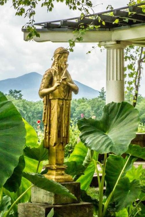 A golden statue of a woman stands among large green leaves, with mountains and a cloudy sky in the background.