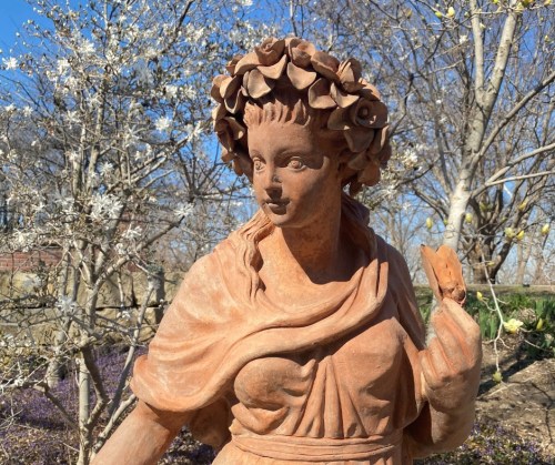 A terracotta statue of a woman with a floral crown, holding a flower, surrounded by blooming trees.