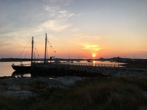 A serene sunset over a harbor with a sailboat and wooden pier, surrounded by calm waters and distant land.