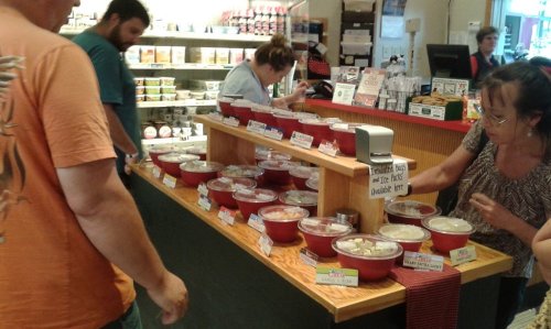 A busy deli counter with various food samples in red bowls, customers sampling and browsing the offerings.