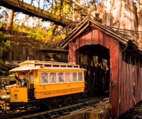 A miniature yellow train passes through a red covered bridge in a scenic outdoor setting.
