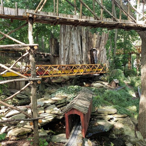 A whimsical wooden bridge connects treehouses in a lush, green forest setting with a small stream below.