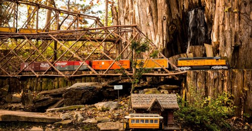 A miniature train set runs through a forest scene with a wooden bridge, rocks, and a small house.