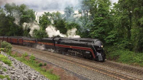 A vintage steam locomotive chugs along a track, surrounded by lush greenery and emitting plumes of smoke.