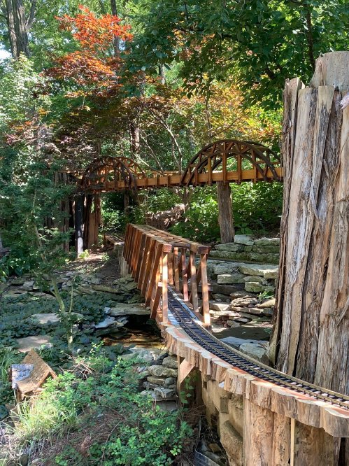 A wooden miniature train track winds through a lush green landscape with trees and rocks.