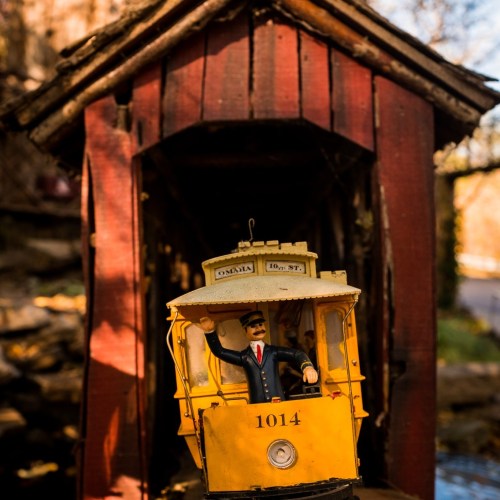 A vintage toy tram with a conductor figure emerges from a rustic wooden bridge surrounded by autumn foliage.