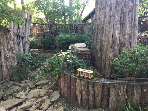 A miniature train travels along a winding track through a lush garden with large trees and rustic wooden structures.