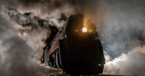 A vintage steam locomotive emerges from fog, illuminated by its headlights against a dark, smoky background.