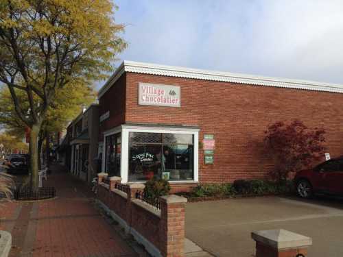 A brick building with a sign reading "Village Chocolatier," surrounded by trees and a sidewalk.