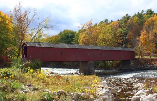 A red covered bridge spans a river, surrounded by vibrant autumn foliage and rocky riverbanks.