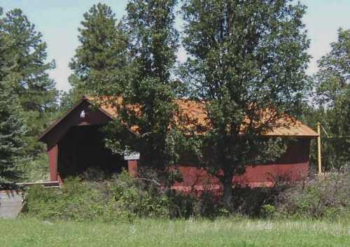 A red covered bridge partially obscured by trees, with a rust-colored roof and surrounded by greenery.