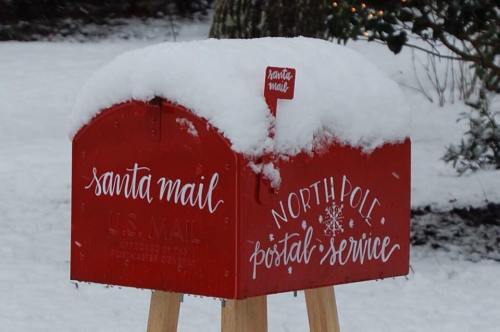 A red mailbox labeled "Santa Mail" covered in snow, standing on a wooden post in a wintery landscape.