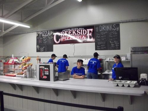 Staff members in blue shirts work behind the counter of Creekside Pizzeria, with a menu board and food displays visible.